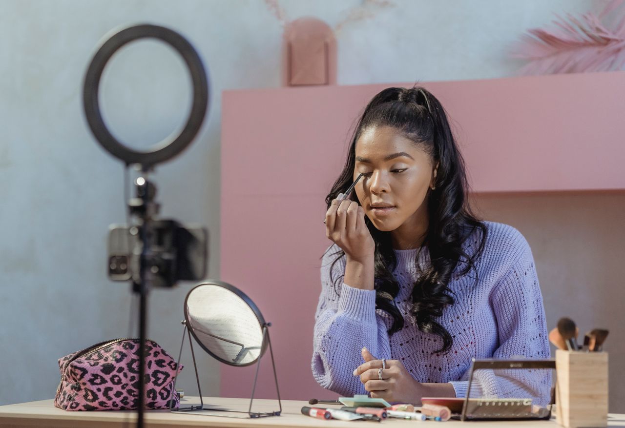 Concentrated African American woman making makeup while filming beauty blog on smartphone in stylish room